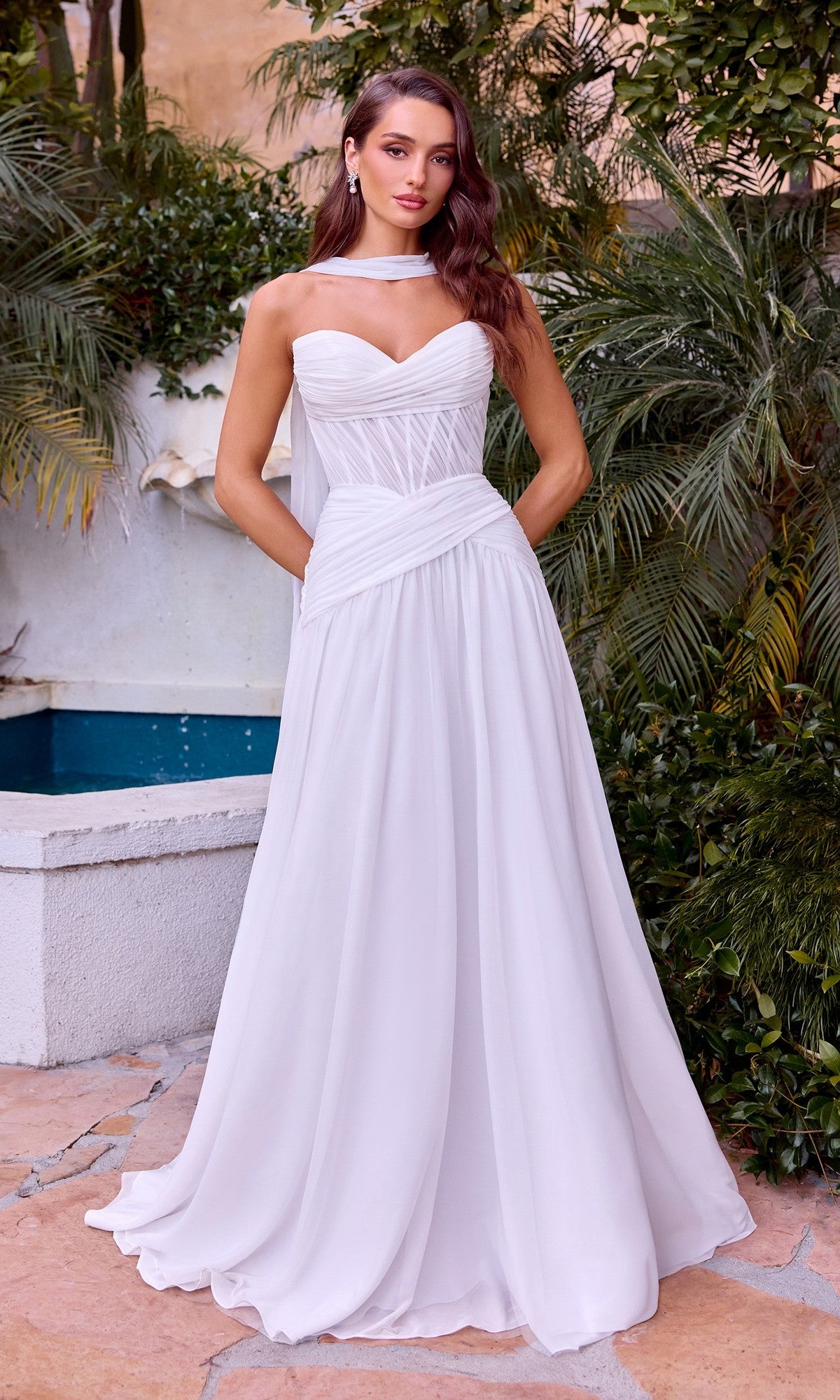 A woman stands outdoors in the Off-White A-Line Prom Dress: Ladivine CD395W, featuring a sheer corset bodice, ruched details, and matching neck scarf, surrounded by greenery and a small pool.